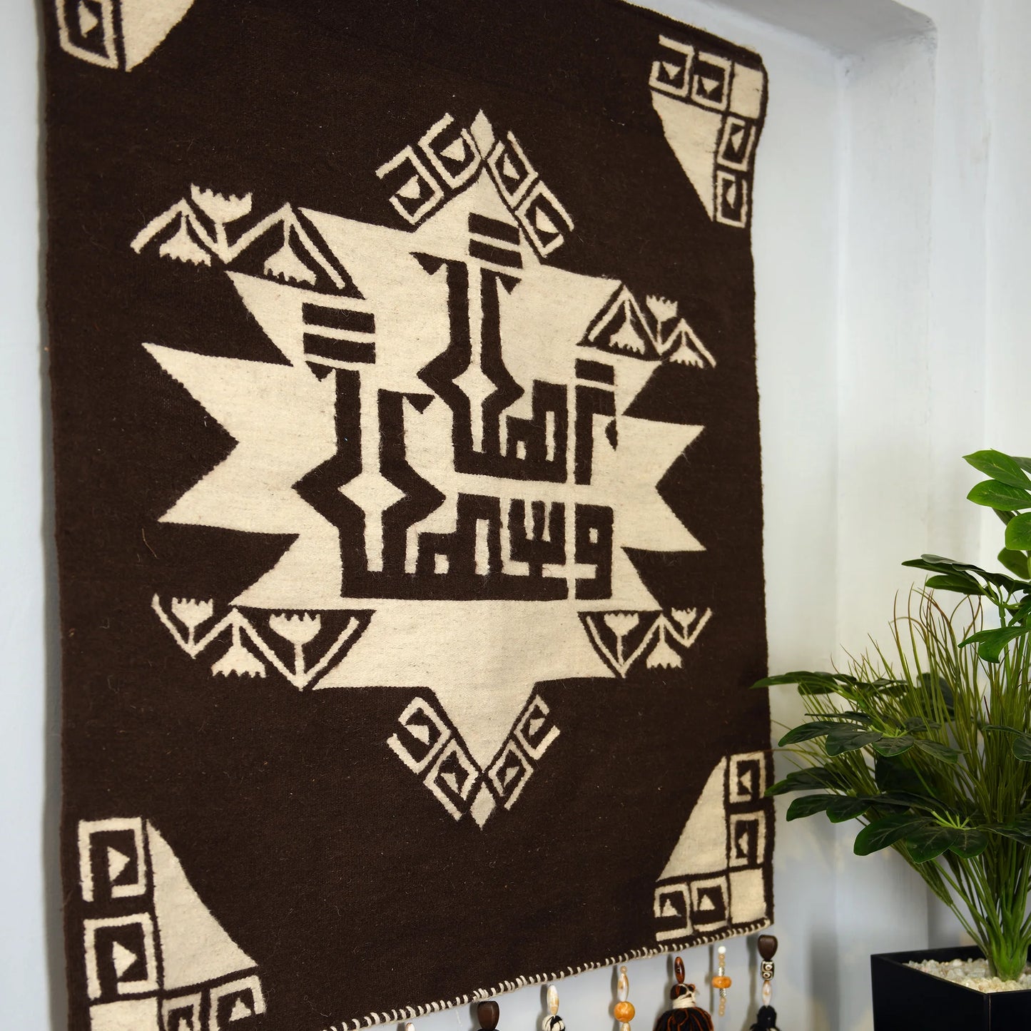 "A detailed view of a woven tapestry with a Brown and Beige geometric pattern hanging on a wall above a brown sofa. The tapestry has a central design with symmetrical shapes and decorative tassels at the bottom."