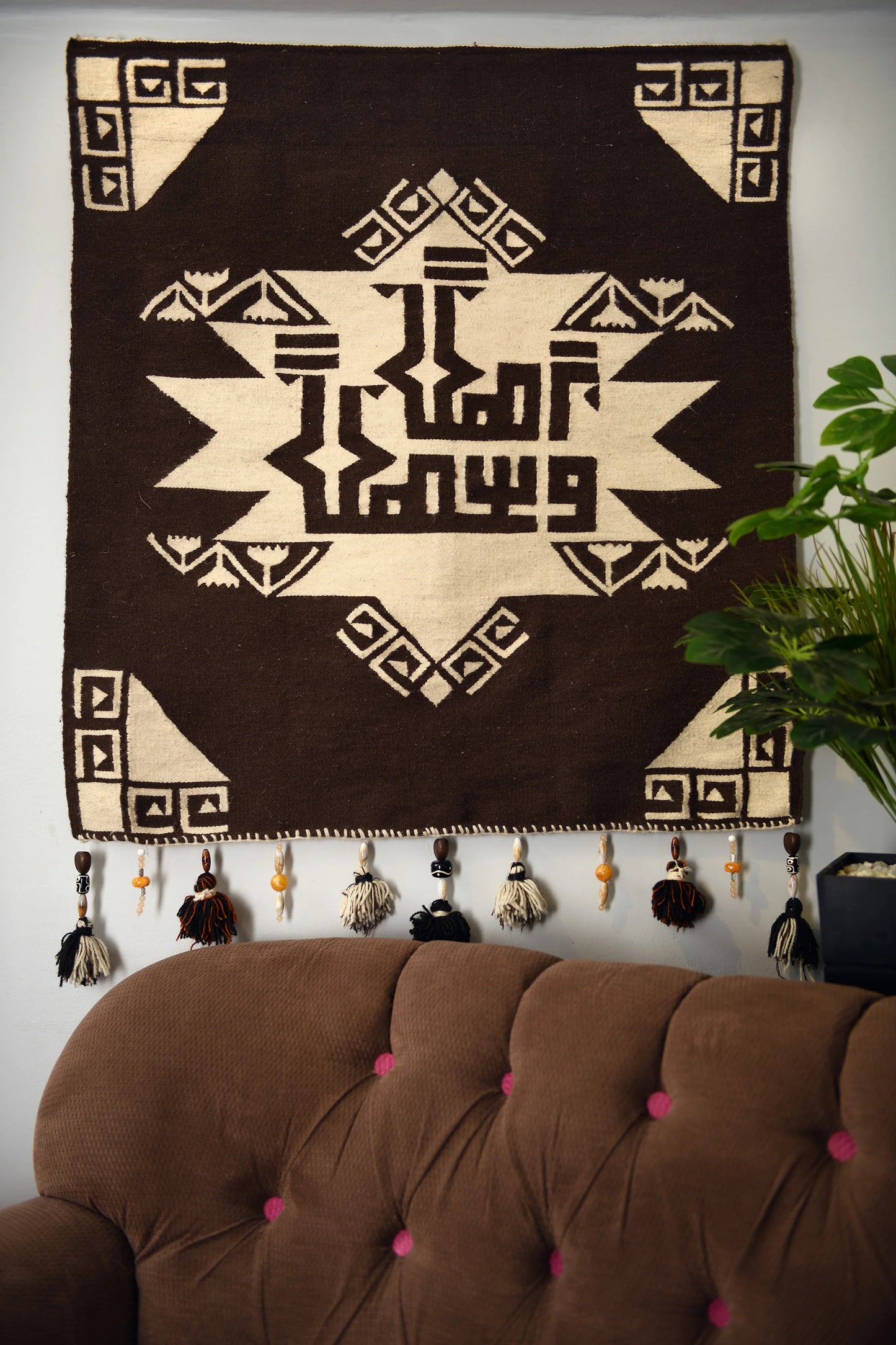 "A close-up of a woven tapestry with a brown and beige geometric pattern hanging on a wall. The tapestry features a central design with symmetrical shapes and decorative tassels at the bottom, with a potted plant visible on the right side."