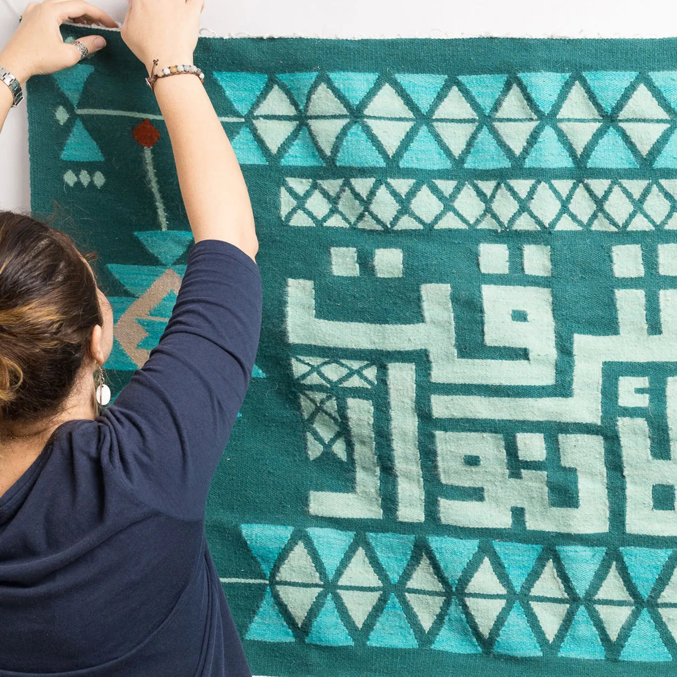 "A woman pointing at a decorative wall hanging with Arabic calligraphy and geometric patterns in green and mint, with her back to the camera."