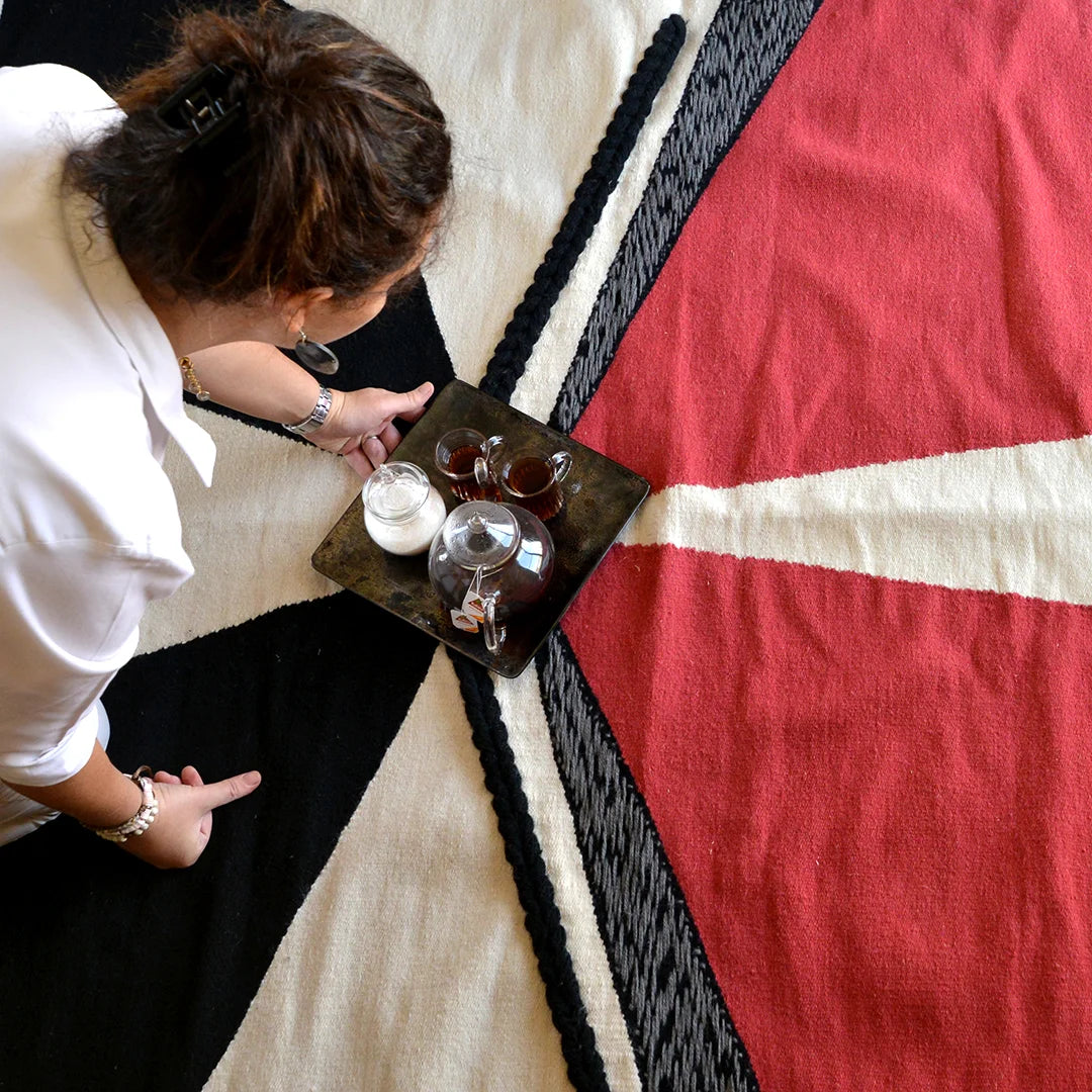 A person is seen from above, placing a tray with glass cups and a teapot on a colorful woven rug. The rug features bold geometric patterns in red, black, and white. The individual is wearing a white shirt and appears focused on arranging the items on the tray.