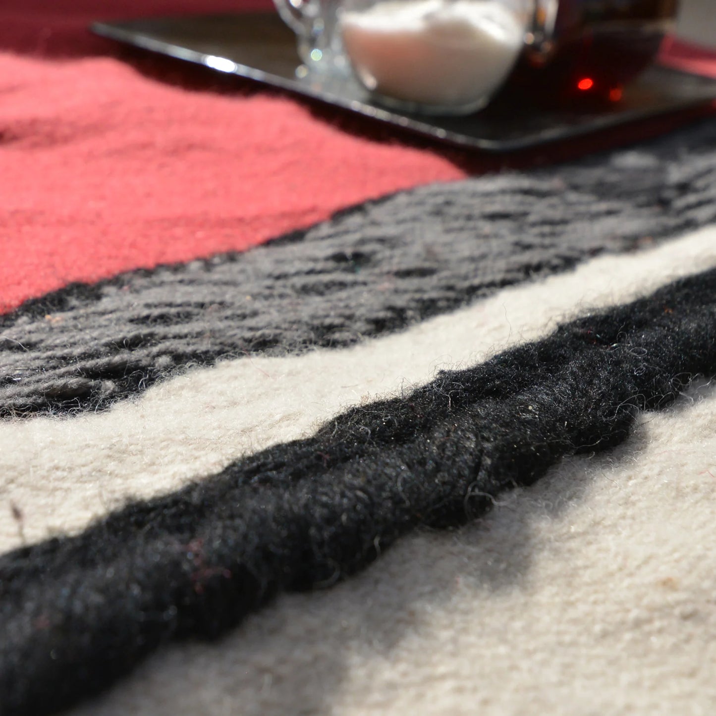 A close-up view of a textured fabric featuring a pattern with black, white, gray, and red stripes. In the background, there is a tray holding a glass container with sugar and a dark liquid, possibly tea or coffee. The focus is on the fabric's fibers and patterns.