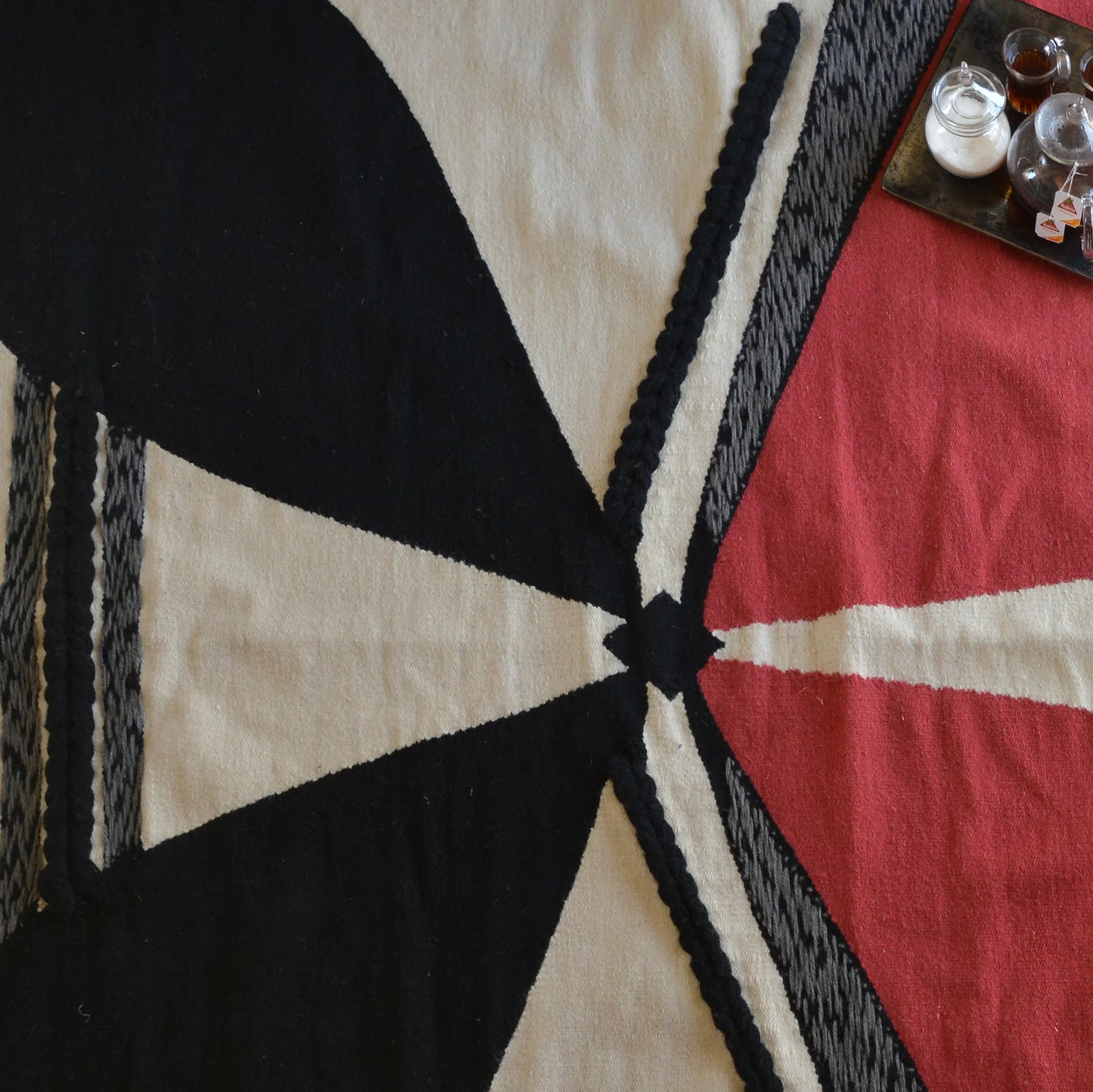 The image features a textile or blanket with a bold geometric design. It includes large sections of black, red, and cream colors, arranged in an abstract pattern with triangular shapes and lines. There are also some small containers or bowls on a tray in the upper right corner, possibly containing spices or other items. The overall composition highlights the contrast between the colors and the intricate design of the fabric.