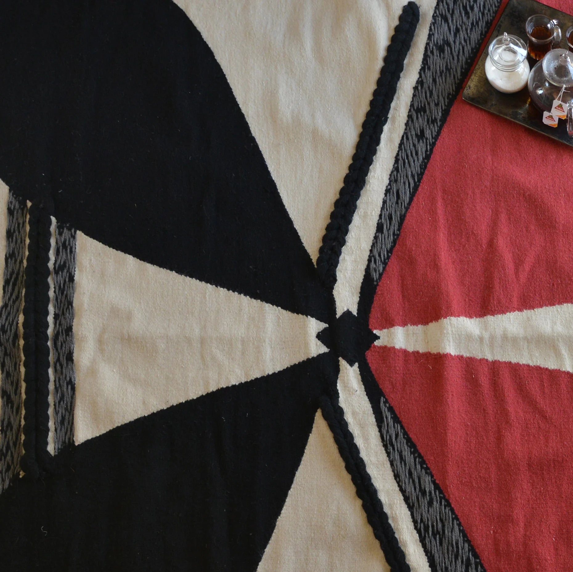 The image features a textile or blanket with a bold geometric design. It includes large sections of black, red, and cream colors, arranged in an abstract pattern with triangular shapes and lines. There are also some small containers or bowls on a tray in the upper right corner, possibly containing spices or other items. The overall composition highlights the contrast between the colors and the intricate design of the fabric.