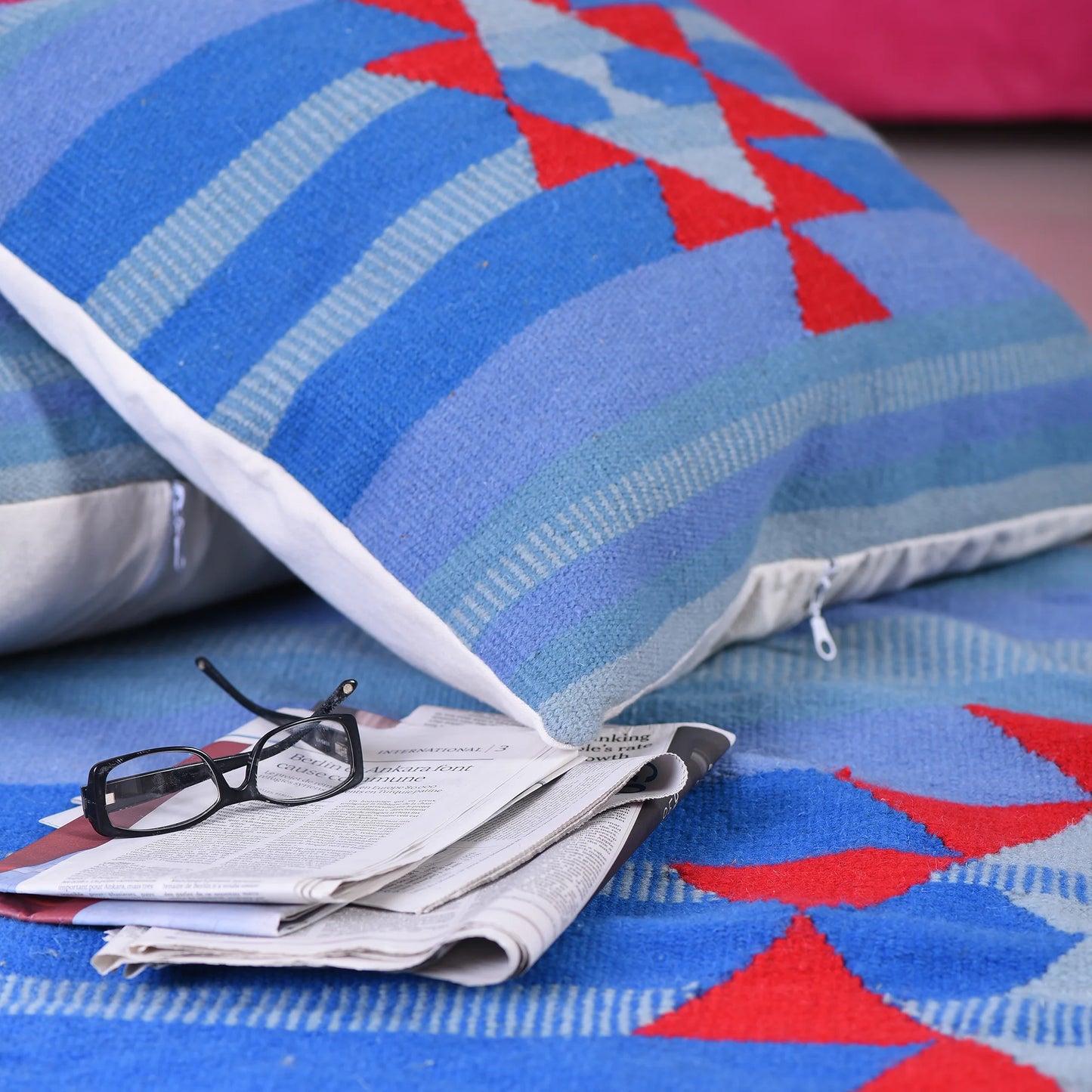 "Two decorative blue and red cushions on a patterned blue rug, with a pair of black glasses resting on a stack of newspapers."