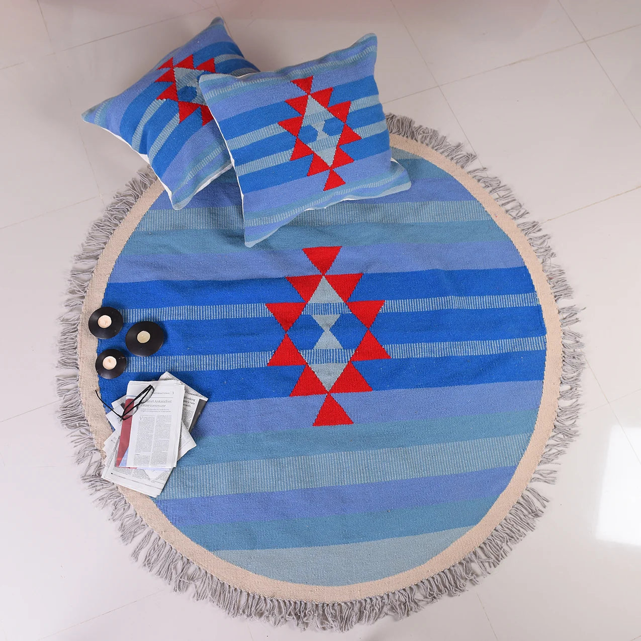 A round, blue area rug with a geometric pattern featuring red triangles arranged in a diamond shape. Surrounding the rug are two square pillows that match the rug's design. There are also three small black coasters and some magazines placed on the rug, set against a light-colored floor.