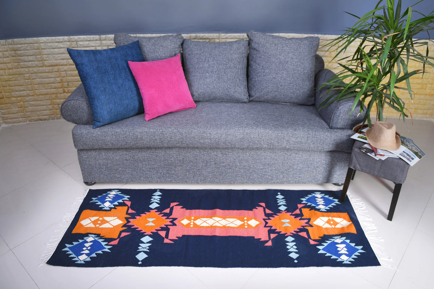 A gray couch adorned with two decorative pillows—one blue and one pink—sits in a stylish interior. In front of the couch, a colorful patterned rug features shades of orange, blue, and white. A small side table next to the couch holds a hat and some magazines, while a green plant adds a touch of nature to the scene.
