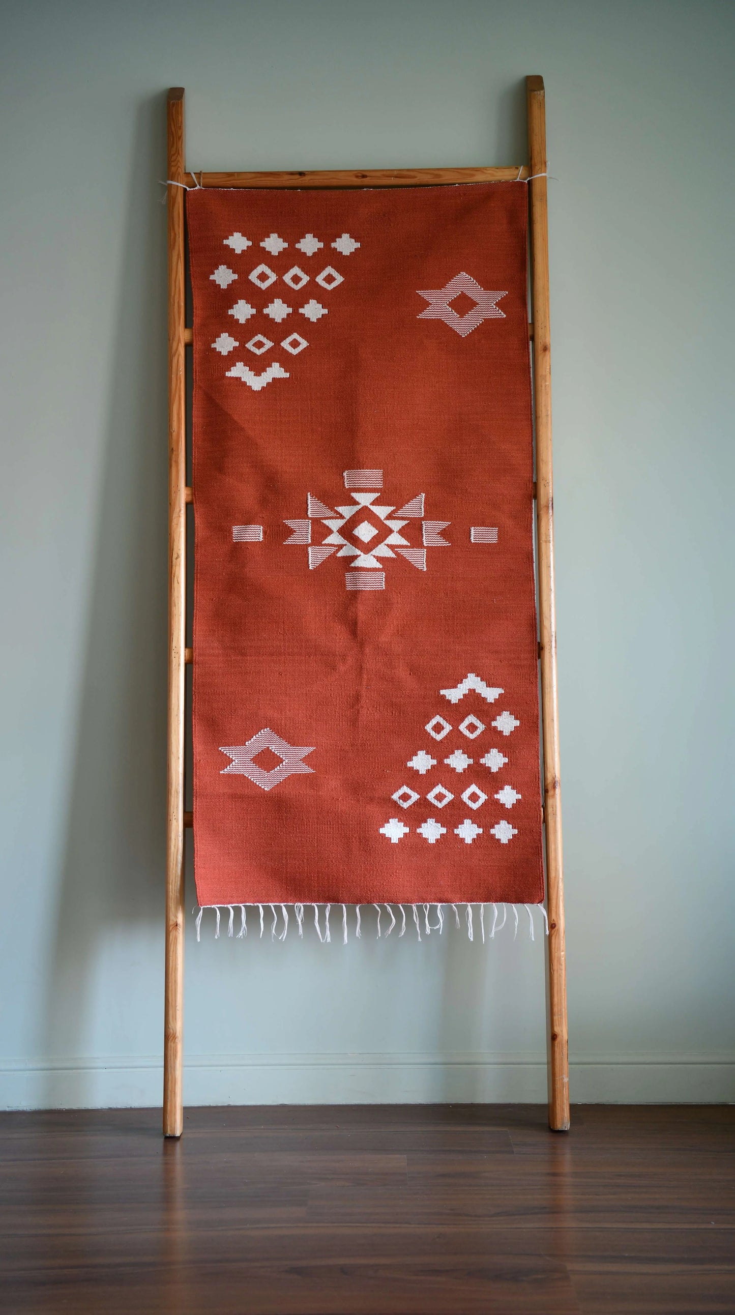 A woven wall hanging is displayed on a wooden ladder against a light green wall. The textile features a rich terracotta background adorned with geometric white patterns, including diamonds and crosses. The design showcases a blend of modern and traditional aesthetics, while the fringed bottom adds a touch of texture to the piece. The overall composition creates a warm and inviting atmosphere.