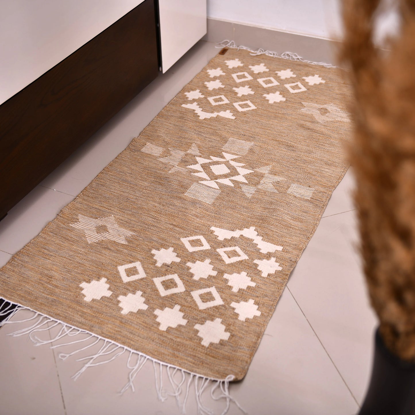 A woven rug is placed on a tiled floor near a low wooden cabinet. The rug features a warm beige background adorned with white geometric patterns, including diamonds and crosses. The fringed edges add texture, and the soft lighting creates a cozy atmosphere. In the foreground, dried plants in a vase enhance the decor, complementing the rug's design.
