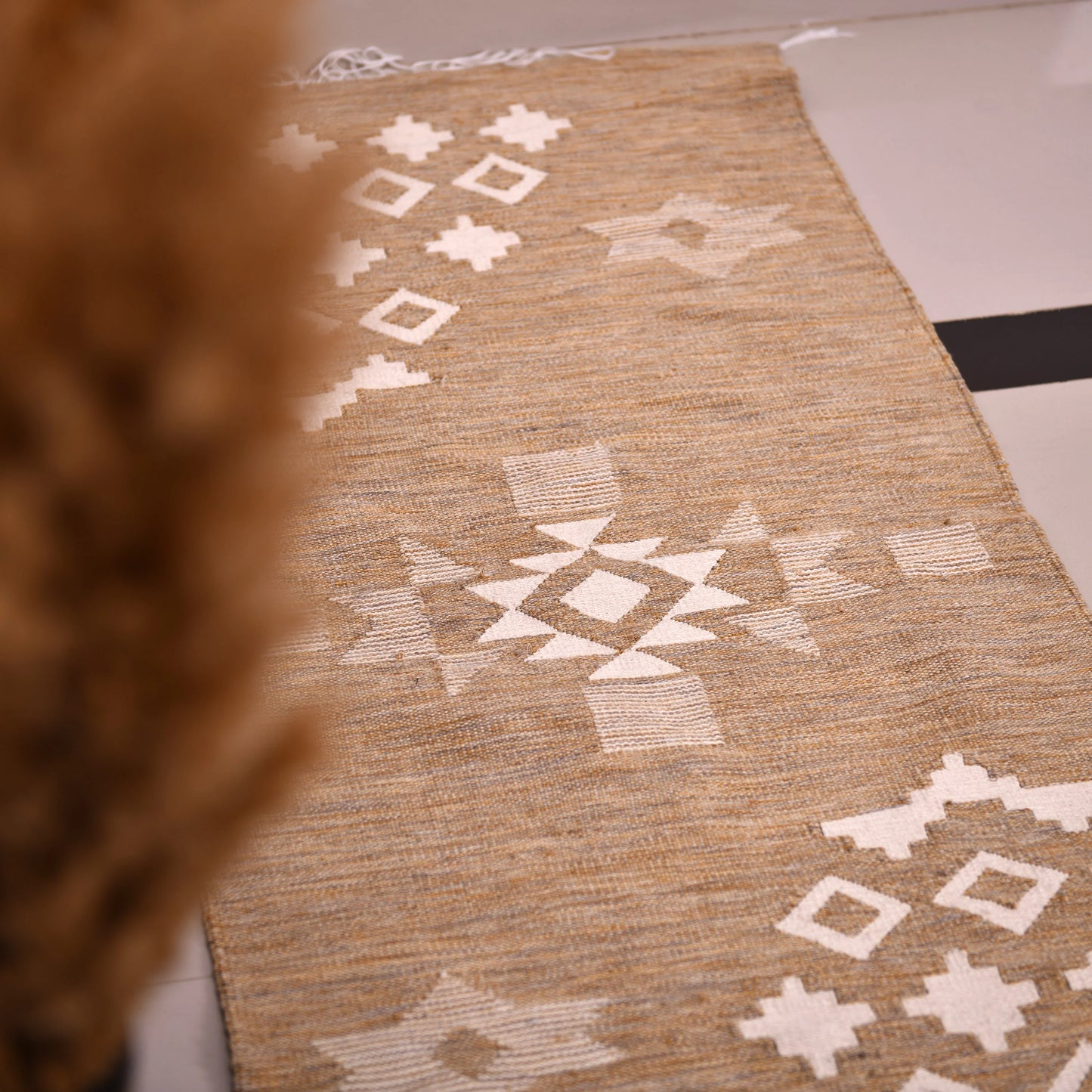 A woven rug is placed on a tiled floor near a low wooden cabinet. The rug features a warm beige background adorned with white geometric patterns, including diamonds and crosses. The fringed edges add texture, and the soft lighting creates a cozy atmosphere. In the foreground, dried plants in a vase enhance the decor, complementing the rug's design.