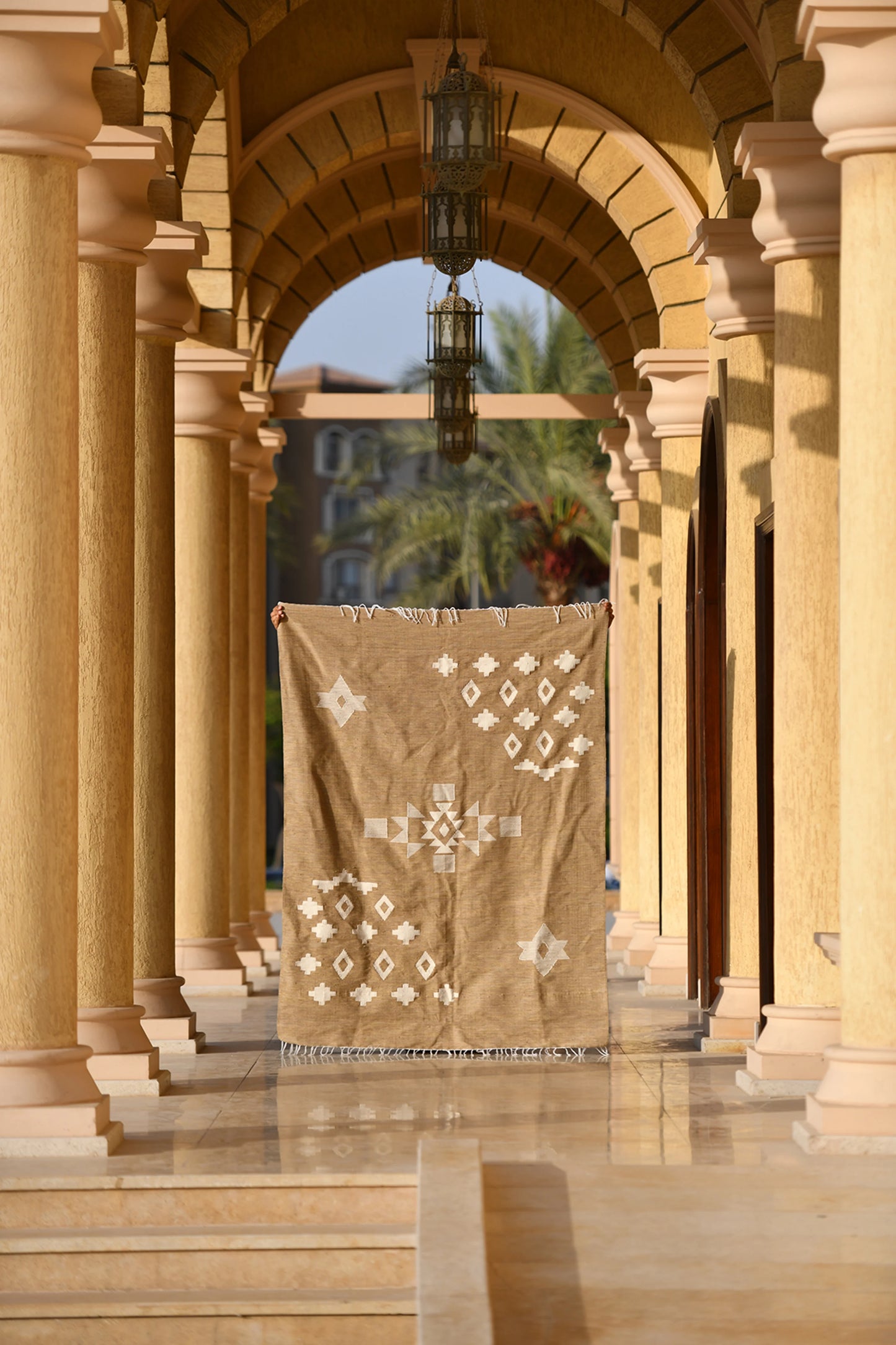 A large woven rug is displayed hanging in an archway, showcasing its warm beige color and white geometric patterns, including diamonds and crosses. The setting features elegant stone columns and a polished floor, bathed in soft sunlight. Palm trees are visible in the background, adding a tropical touch to the serene architectural scene. The combination of the rug and surroundings creates a harmonious blend of texture and elegance.