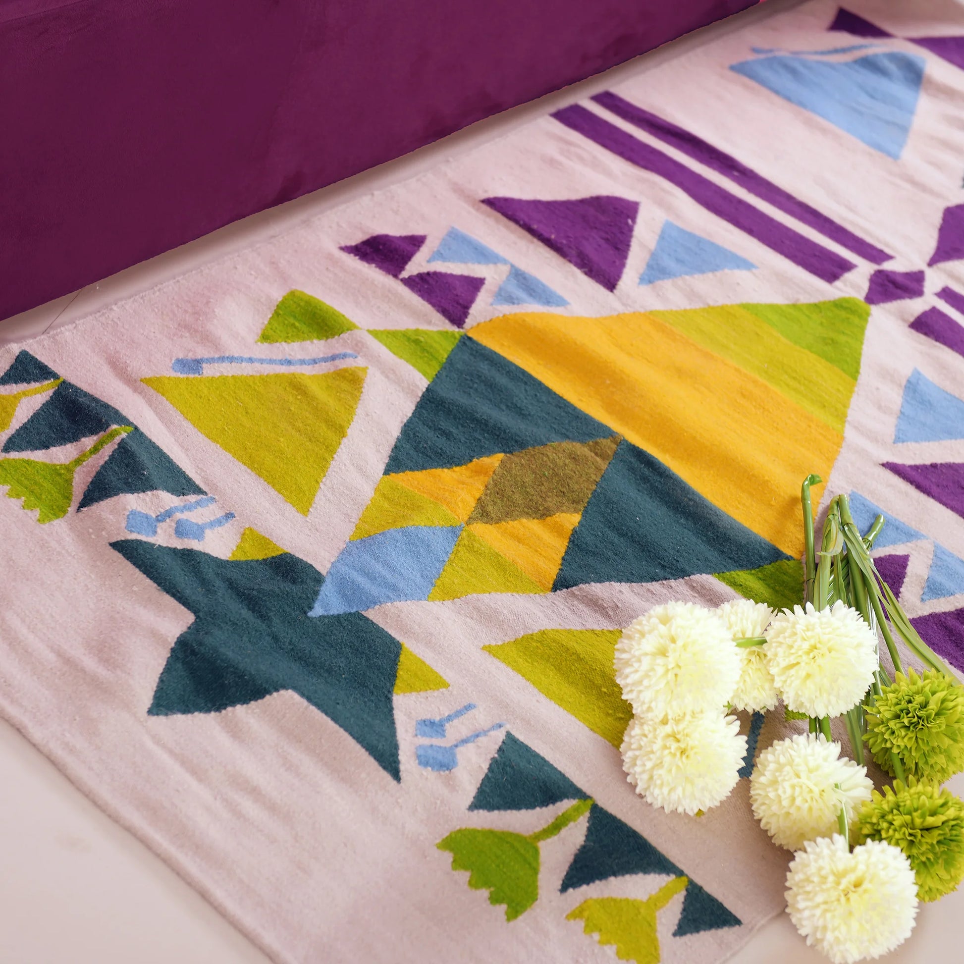"A close-up of a woven rug with geometric patterns in green, yellow, blue, and purple on a light beige background, with a bouquet of white and green flowers placed on it."