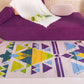 "A woven rug with geometric patterns in green, yellow, blue, and purple on a light beige background, placed on a floor in front of a purple couch with a white blanket and flowers."