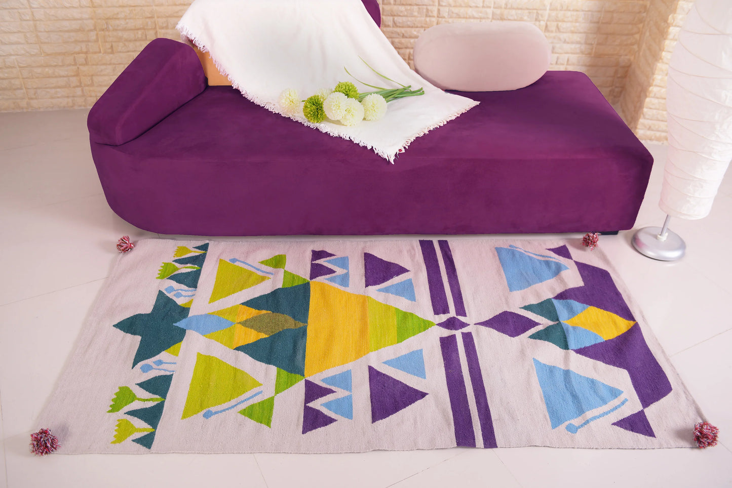 "A woven rug with geometric patterns in green, yellow, blue, and purple on a light beige background, placed on a floor in front of a purple couch with a white blanket and flowers."