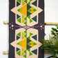 "A vertically displayed woven rug with geometric patterns in green, yellow, blue, and purple on a dark background, hung on a bamboo rod with a plant in the background."