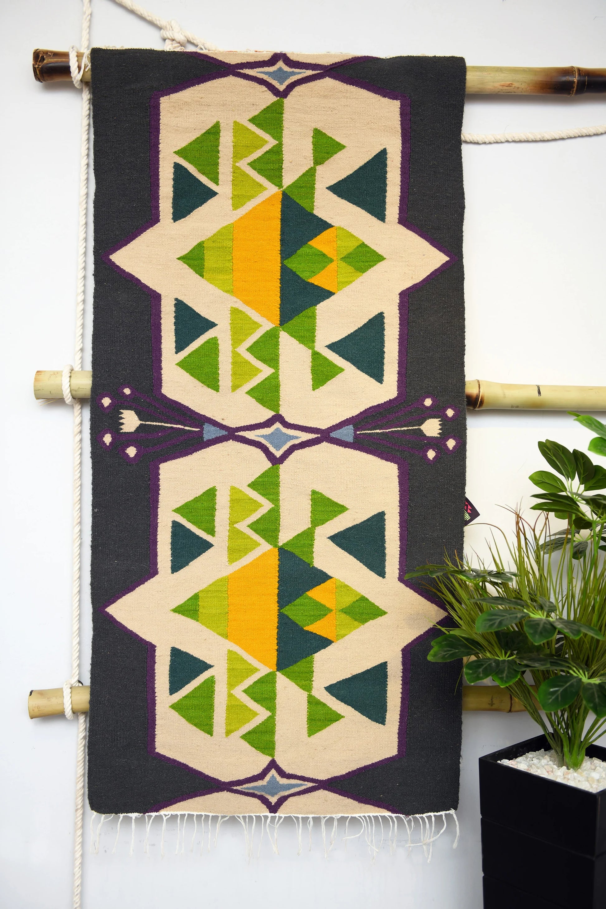 "A vertically displayed woven rug with geometric patterns in green, yellow, blue, and purple on a dark background, hung on a bamboo rod with a plant in the background."