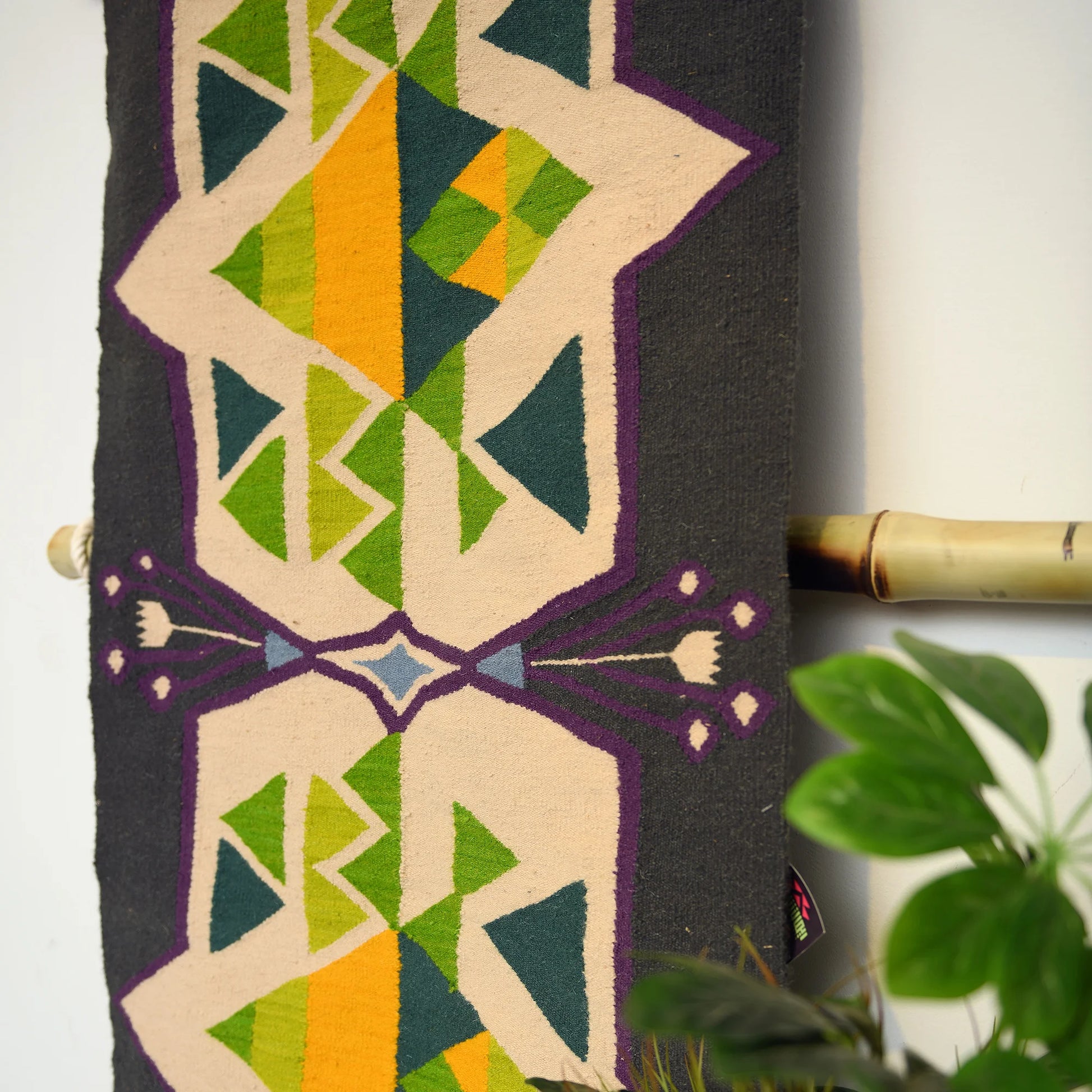 "A close-up of a vertically displayed woven rug with geometric patterns in green, yellow, blue, and purple on a dark background, hung on a bamboo rod."
