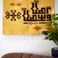 "A close-up of a woven tapestry with a yellow background featuring brown geometric patterns and Arabic calligraphy, hanging on a wall. The tapestry has decorative fringes at the bottom, with a potted plant visible on the right side."