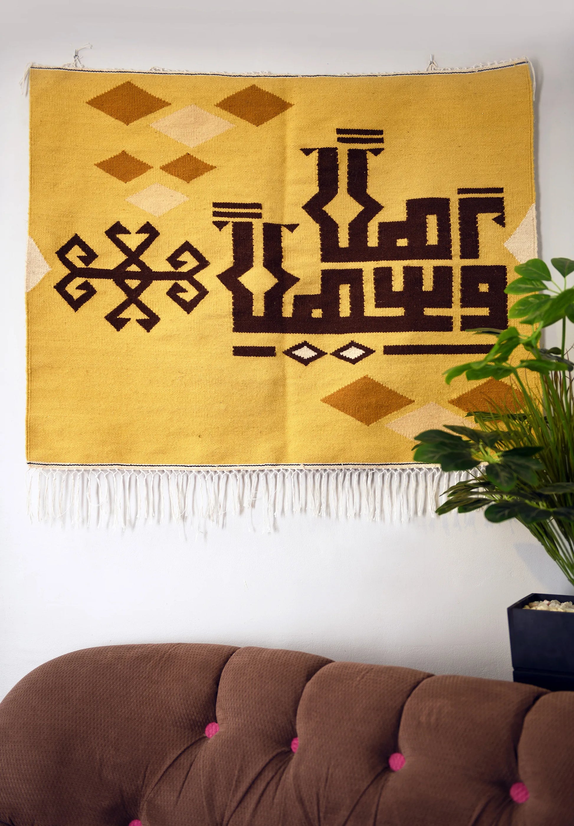 "A close-up of a woven tapestry with a yellow background featuring brown geometric patterns and Arabic calligraphy, hanging on a wall. The tapestry has decorative fringes at the bottom, with a potted plant visible on the right side."