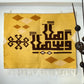 "A detailed view of a woven tapestry with a yellow background featuring brown geometric patterns and Arabic calligraphy, hanging on a wall above a brown sofa. The tapestry has decorative fringes at the bottom."