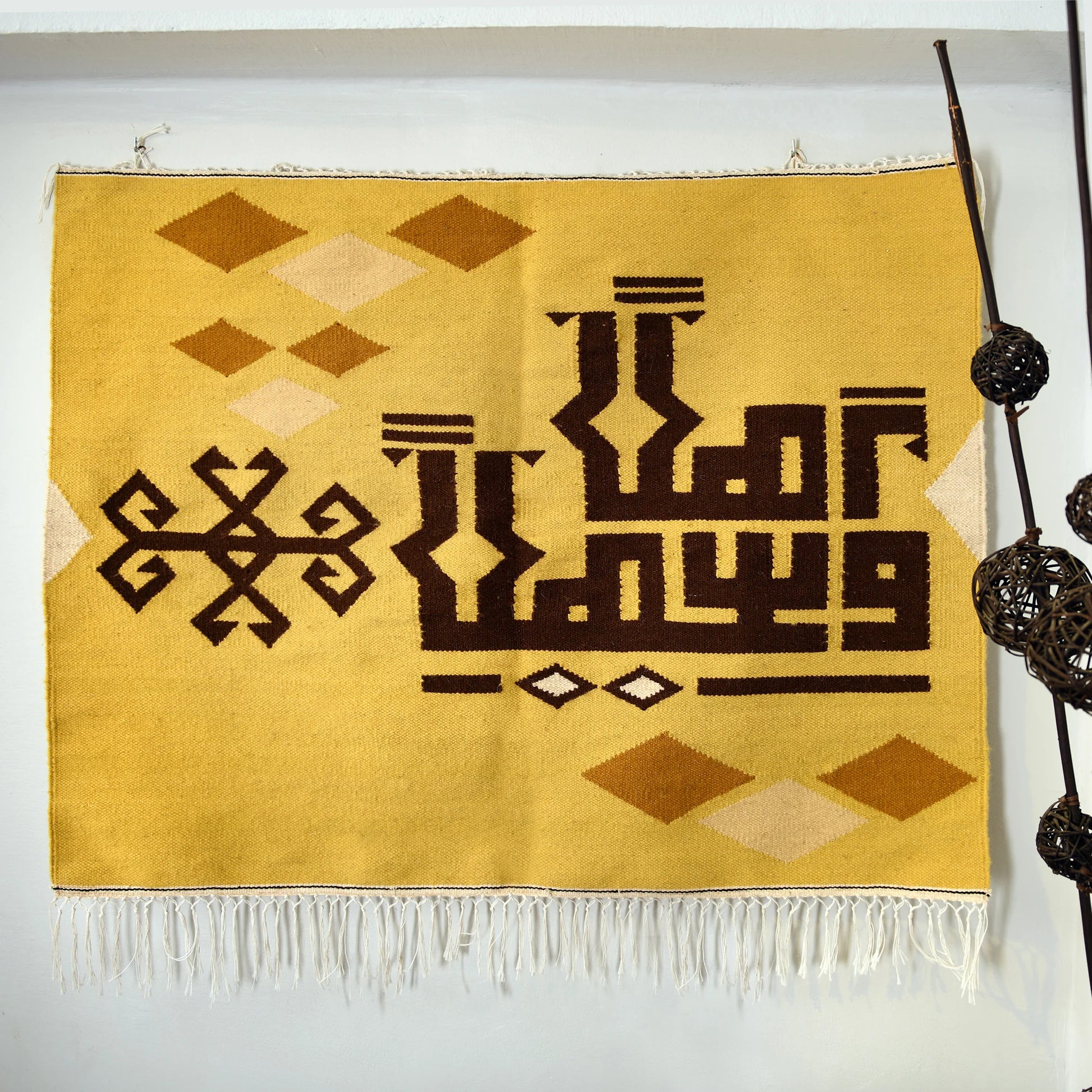 "A detailed view of a woven tapestry with a yellow background featuring brown geometric patterns and Arabic calligraphy, hanging on a wall above a brown sofa. The tapestry has decorative fringes at the bottom."