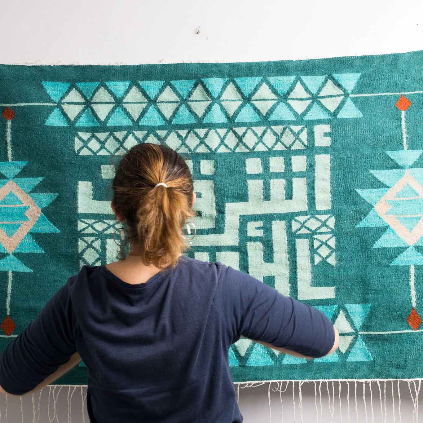"A woman with her back to the camera, looking at a decorative wall hanging with Arabic calligraphy and geometric patterns in green and mint."