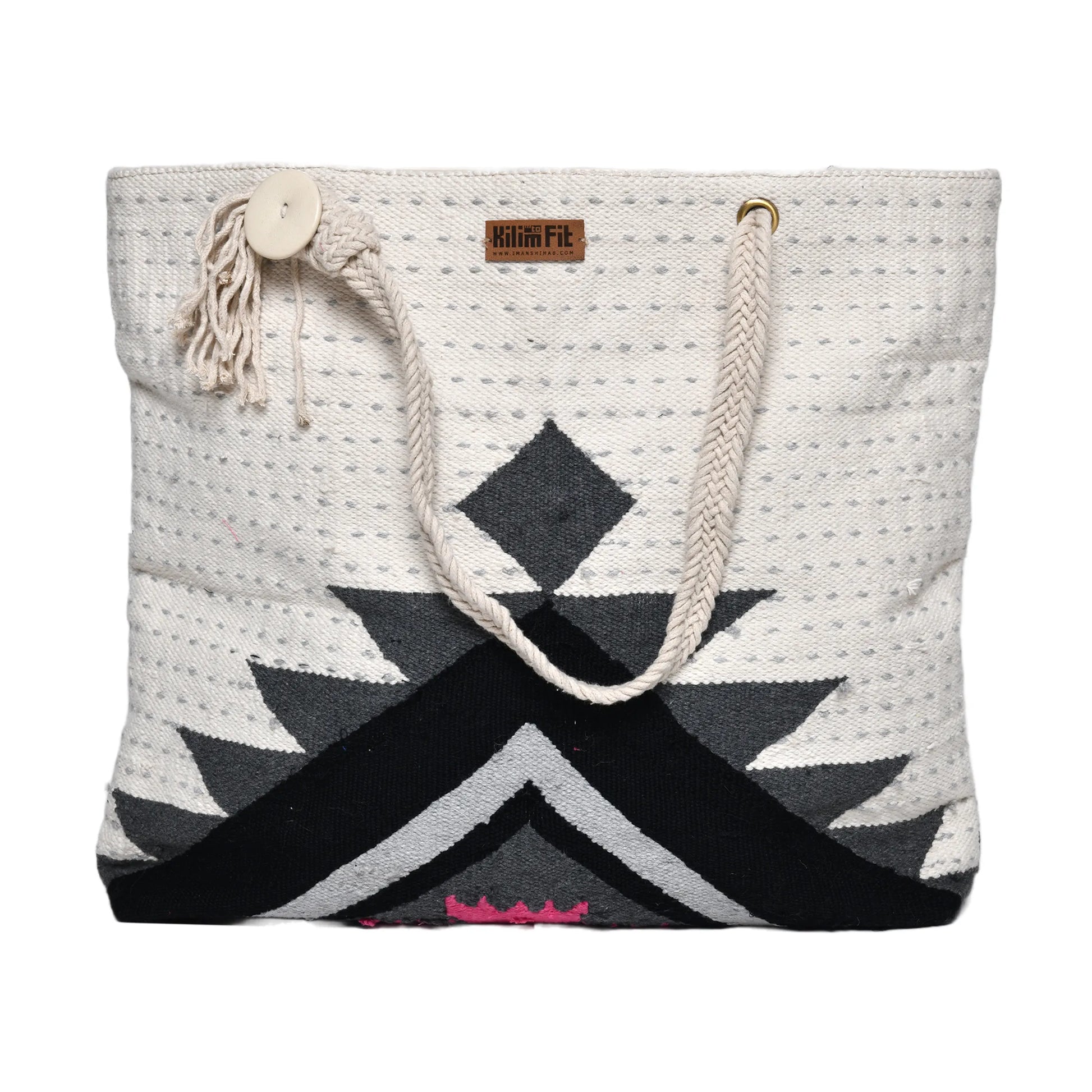 "Front view of a handwoven tote bag with a geometric pattern in black, gray, white, and pink colors. The bag has rope handles and a button closure with a tassel."