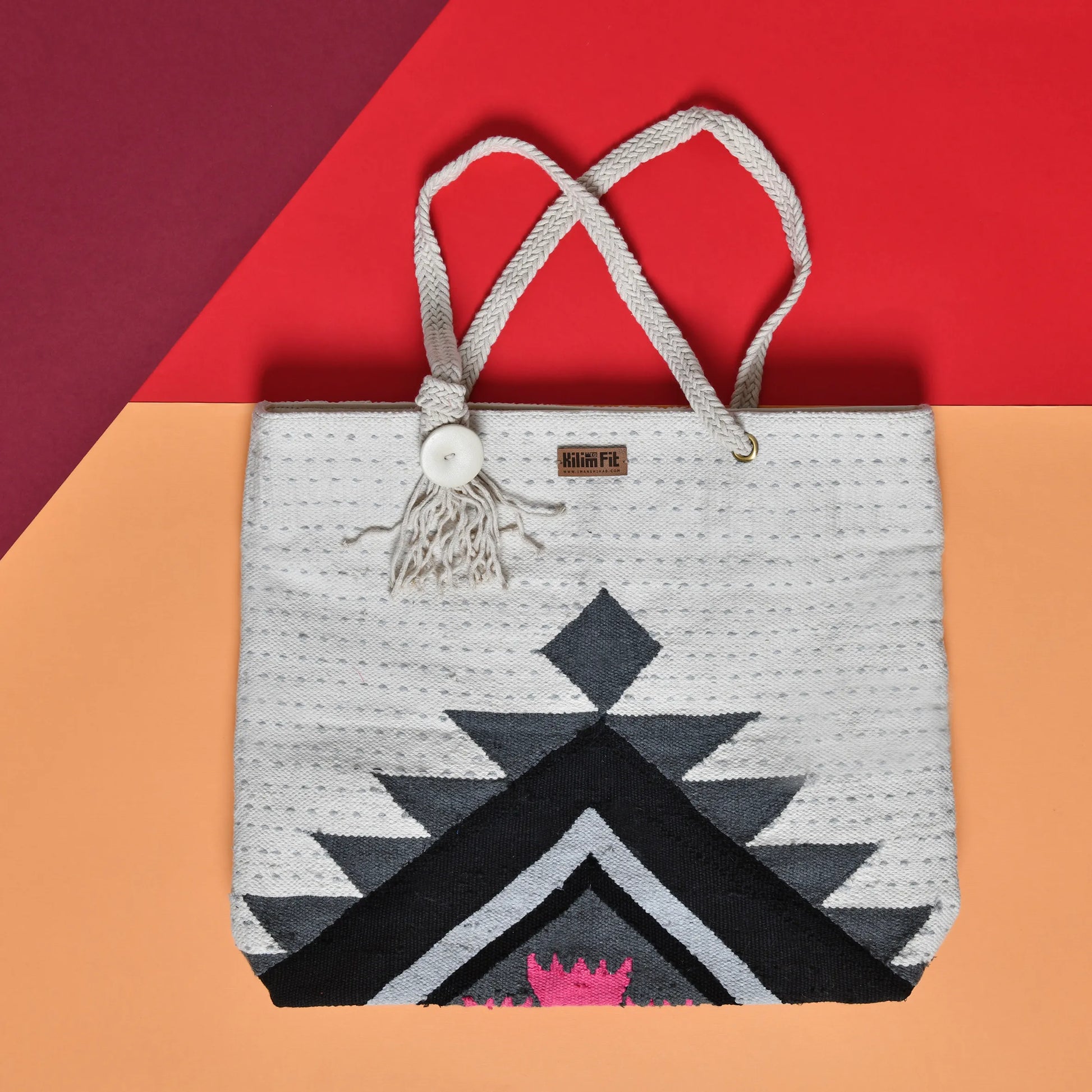 "A woven tote bag with a geometric pattern in black, white, and gray, placed on a colorful background, featuring a rope handle and a tassel."