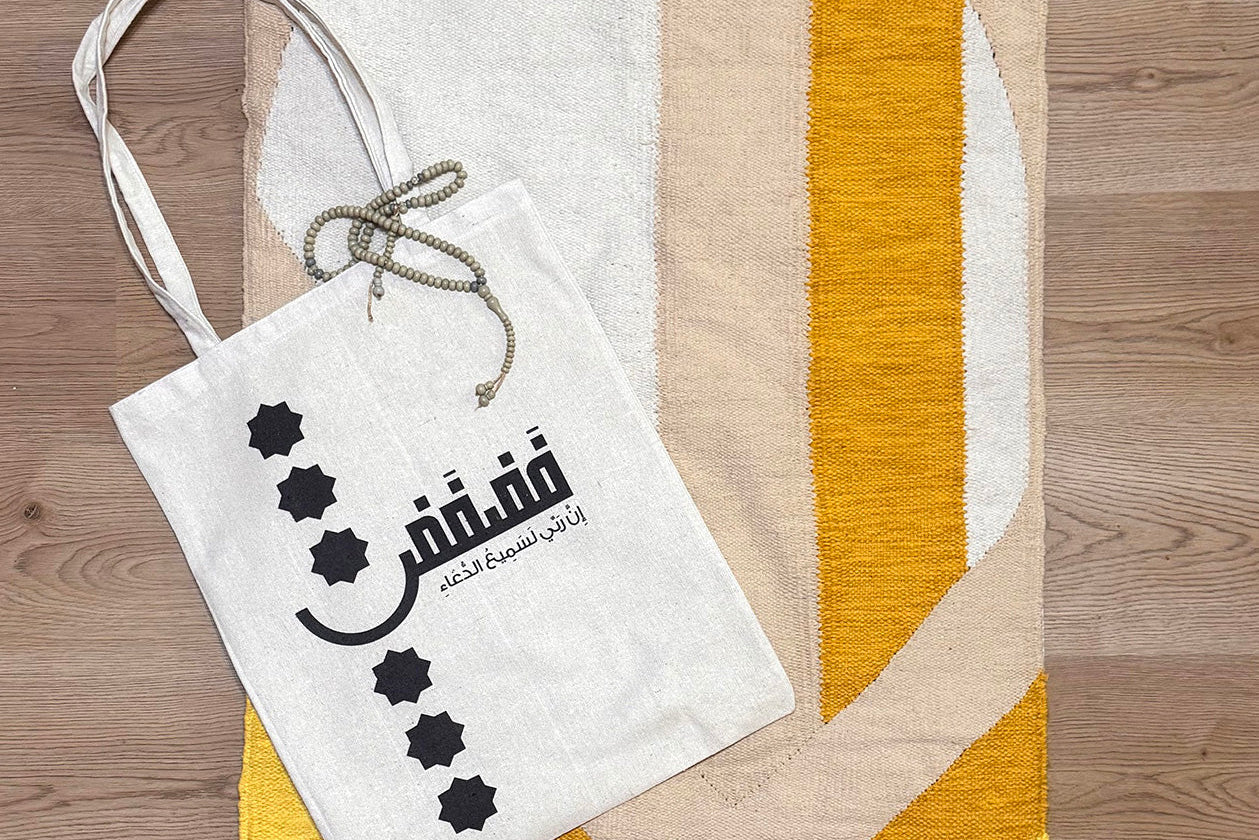 White tote bag with Arabic text and star designs on a wooden surface with a woven mat.