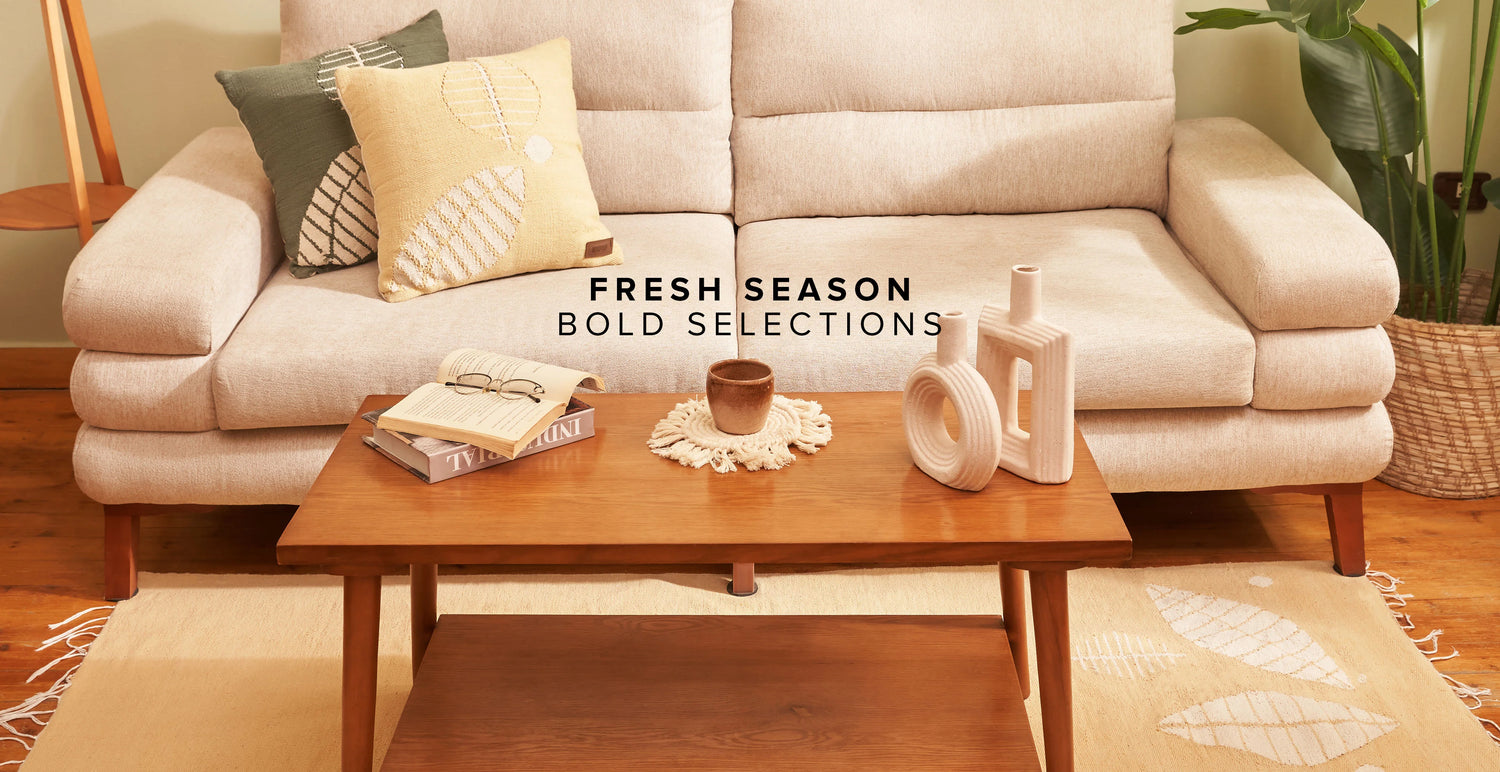 A cozy living room setting featuring a neutral-toned sofa with two decorative pillows, one green and one yellow with geometric patterns. A wooden coffee table in the center holds a small vase, a cup, and a stack of books. A woven rug with subtle leaf patterns rests on the floor, and a potted plant adds a touch of greenery. The text "Fresh Season Bold Selections" is prominently displayed, highlighting the modern and inviting atmosphere of the space.