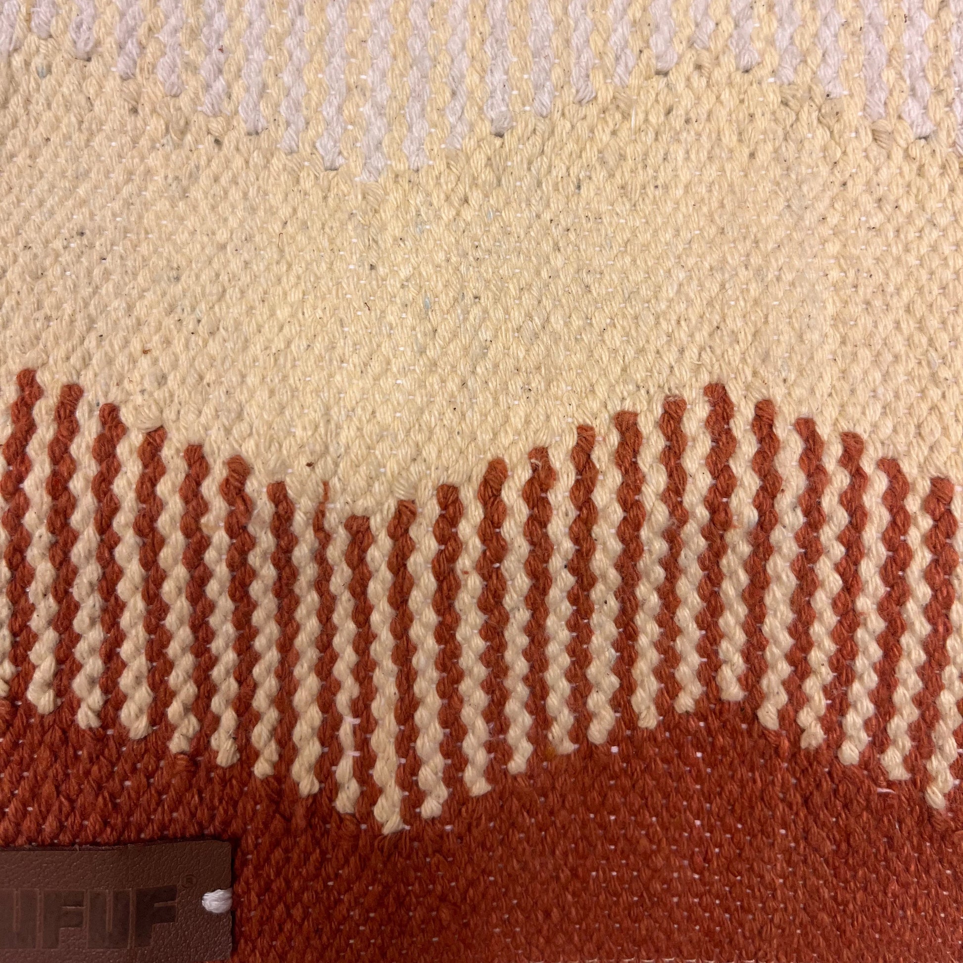 A close-up of woven fabric showcases a textured pattern in beige, cream, and terracotta hues. The design features alternating smooth and ribbed sections, highlighting the craftsmanship. A small brown leather tag is partially visible, adding to the aesthetic.