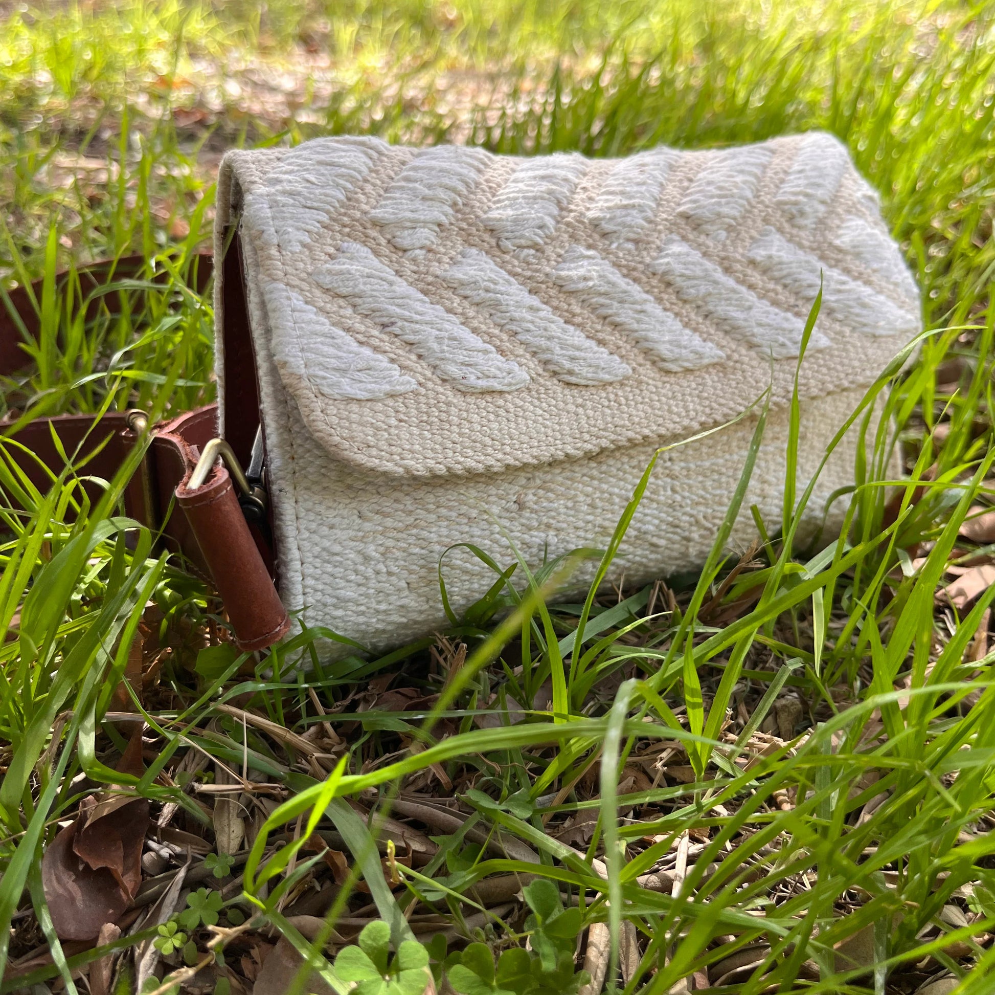 "A cream-colored woven bag with a white chevron pattern, positioned among green grass. The bag has a brown leather strap and displays its design in a natural setting."