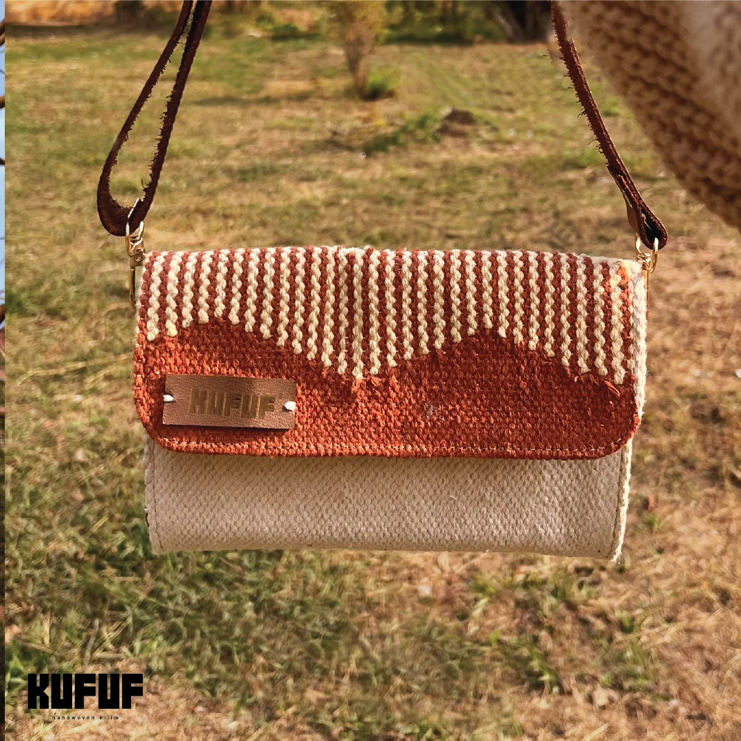 A woven handbag is held against a natural outdoor background, showcasing its terracotta and cream design with a textured pattern. The bag features a brown leather strap and a small metal brand tag that reads "KUFUF." The lush greenery in the background adds a vibrant touch to the scene.