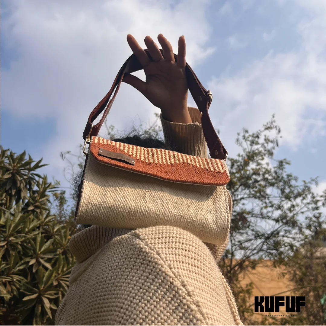 A person holds a woven handbag up against a cloudy sky. The bag features a terracotta and cream design with a textured pattern and a brown leather strap. The person's cozy sweater adds warmth to the composition, while the brand tag "KUFUF" is visible, emphasizing the bag's stylish appeal.